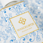 Close-up van het etiket van Wasgeurtje Blossom Drip wasparfum met goud logo op wit-blauw sierpatroon.