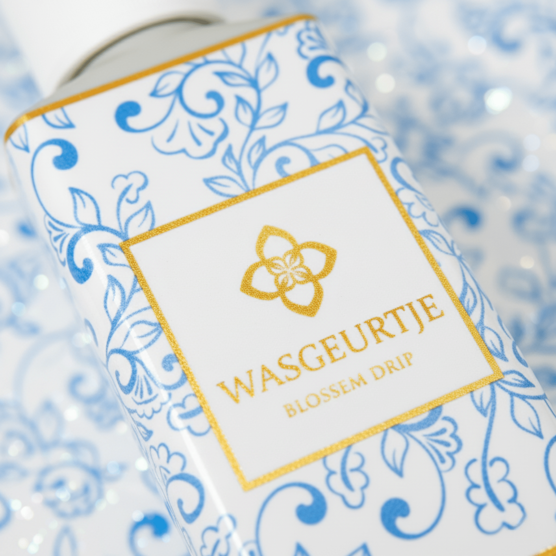 Close-up van het etiket van Wasgeurtje Blossom Drip wasparfum met goud logo op wit-blauw sierpatroon.
