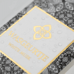 Nauwkeurige close-up van het gouden logo en label van Wasgeurtje White Musk wasparfum met fijne waterdruppels op het oppervlak — een premium detailopname die de luxe afwerking en verfijnde merkidentiteit benadrukt.