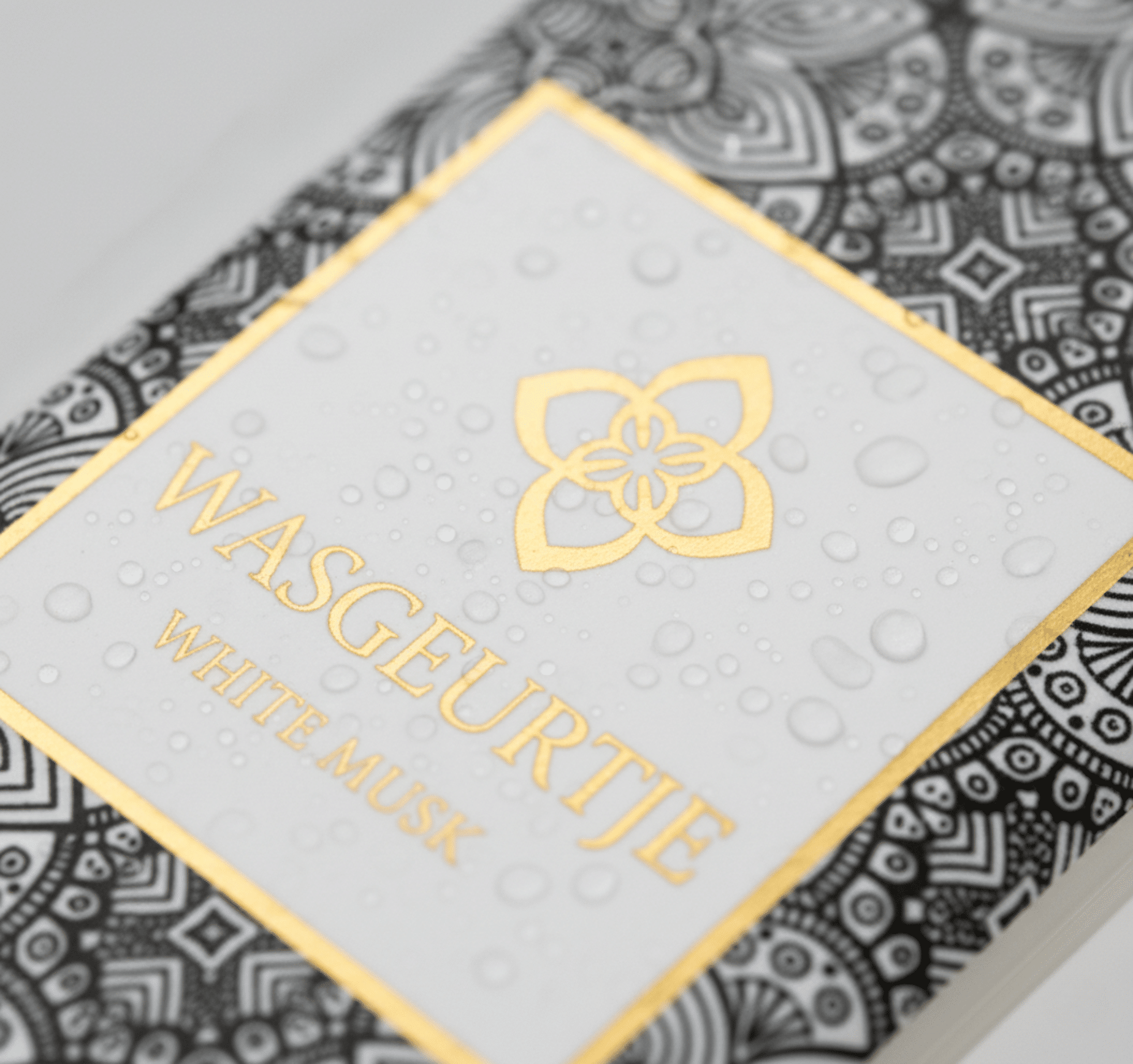 Nauwkeurige close-up van het gouden logo en label van Wasgeurtje White Musk wasparfum met fijne waterdruppels op het oppervlak — een premium detailopname die de luxe afwerking en verfijnde merkidentiteit benadrukt.