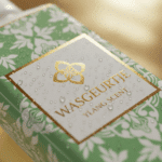 Nauwkeurige close-up van het gouden logo en het label van Wasgeurtje Ylang Scent wasparfum, bedekt met fijne waterdruppels — toont de hoogwaardige afwerking en frisse uitstraling van het product.