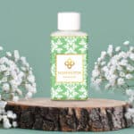 Wasgeurtje Ylang Scent wasmiddel met een groen en wit floraal patroon, naast witte bloesems op een houten schijf.